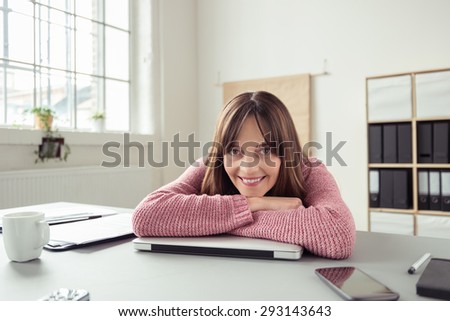 Happy businesswoman taking a break from her work schedule leaning forwards to relax on her folded arms as she smiles happily at the camera
