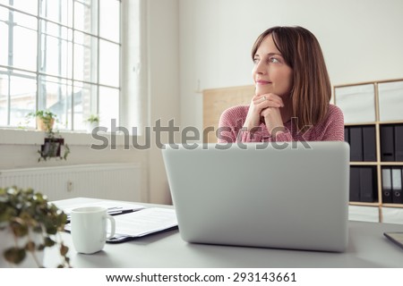 Young woman sitting at her desk in front of a gray laptop while daydreaming of nice memories or future in a modern bright office with large window