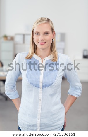 Half Body Shot of a Pretty Young Office Worker Standing Inside the Office with Both Hands on her Hips and Smiling at the Camera.