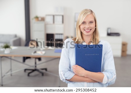 Pretty Blond Young Office Woman Hugging a Blue Documents Folder and Smiling at the Camera Inside the Office.
