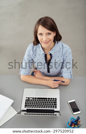 Young Office Woman Sitting at her Desk with Laptop Computer, Smiling at the Camera from High Angle Point.