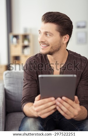 Close up Handsome Young Bearded Guy Holding his Portable Tablet Computer, Looking Into the Distance While Sitting at the Sofa In the Living Room.