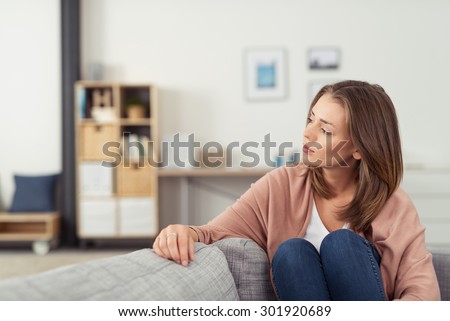 Pensive Young Woman Sitting on a Couch in the Living Room with Knees Up and Looking Into the Distance.