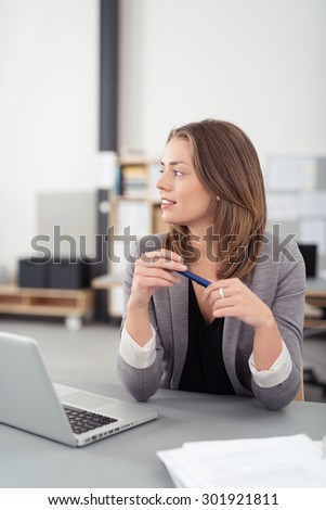 Thoughtful Young Office Woman Sitting at her Table with Laptop and Documents, Looking Into Distance at the Left of the Frame While Holding a Pen.