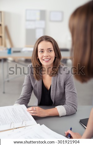 Pretty Young Office Woman Sitting at her Desk with Documents, Listening to her Co-Worker Talking to Her.