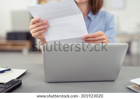 Businesswoman reading a handheld letter or document as she sits at her desk in the office in front of a laptop computer