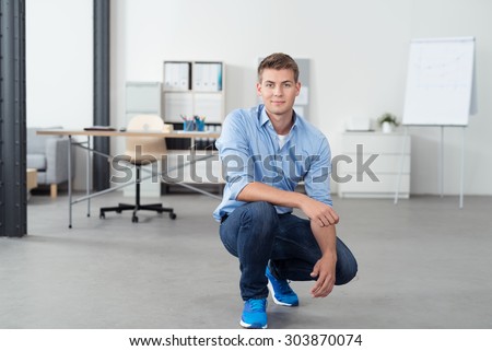 Smiling Handsome Young Office Man Squatting on the Floor Inside his Workplace and Looking at the Camera.