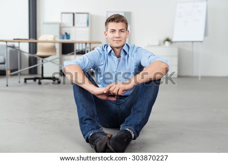 Front View of a Handsome Young Office Guy Sitting on the Floor with Arms Leaning on his Knees and Smiling at the Camera