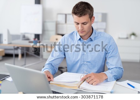 Handsome Young Businessman Sitting at his Desk Inside the Office, Reading Some Documents on a Binder Seriously