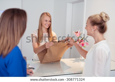 Smiling attractive young shop assistant serving customers in a store handing over a brown paper packet over the counter with a lovely smile