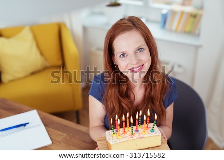 Attractive happy woman celebrating her birthday sitting at the dining table holding a cake with burning candles and looking up at the camera with a lovely smile of pleasure