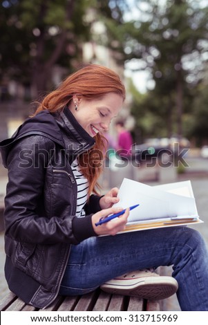 Attractive Young Woman Wearing Casual Winter Outfit, Studying her Lessons at the Outdoor Bench with happy Facial Expression.