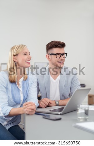 Happy Young Business Couple Sitting at the Table with Laptop Computer and Looking Into the Distance Where Someone is Talking.