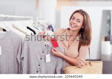 Close up Stylish Female Shopper in a Store Showing the Red Tag of a Gray Shirt at the Camera with Happy Facial Expression.