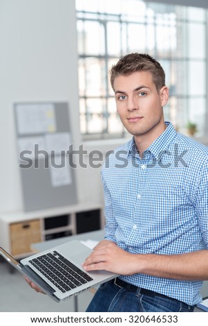 Half Body Shot of a Handsome Young Businessman Holding his Laptop Computer and Looking at Camera Inside the Office.
