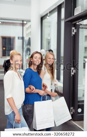Three Young Female Friends Holding White Shopping Bags Inside Retail Clothing Store, Smiling at the Camera.