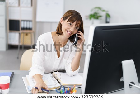 Happy Businesswoman Sitting at her Desk, Talking to Someone on Mobile Phone While Working on her Computer