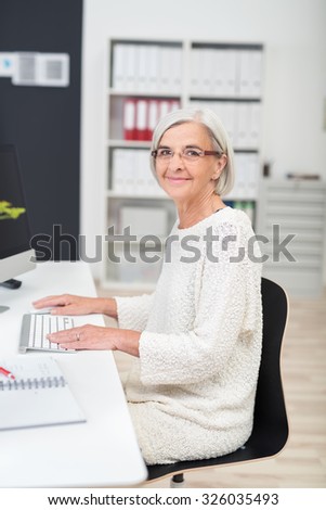 Senior Businesswoman Sitting at her Desk and Smiling at the Camera While Working on her Computer.