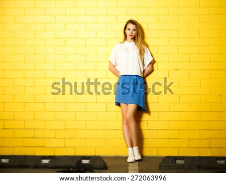 Full Length Portrait of Hipster Fashion Girl with Hands behind her Back Standing at the Yellow Brick Wall Background. Youth Urban Fashion Concept. Copy Space.