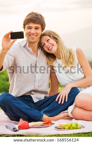 Couple taking photo of themselves with smart phone on romantic picnic date, taking a \