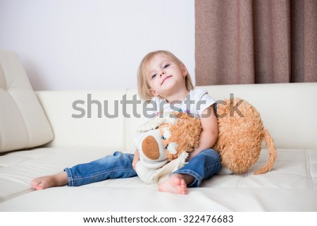 little blonde nice smiling girl three years sitting with a soft toy dog on a white leather couch