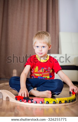 Blonde, blue-eyed little boy of five years in the red T-shirt barefoot beside the couch at home fun playing with wooden railway