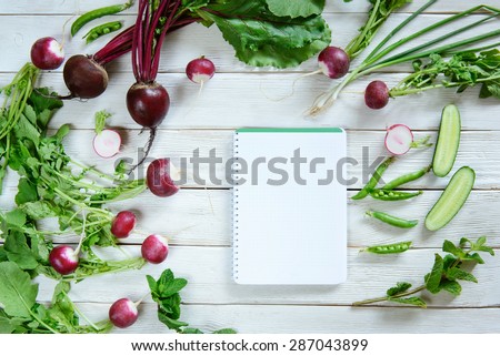 Rural white wood kitchen table with blank cook book for shopping list with salad ingredients (beets, radishes, peas, cucumber, parsley, mint). Blanck recipe book, copy space