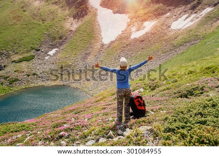 Woman standing on the top of mountain with backpack on a sunlight. Hiker celebrating success. Mountain lake in the valley