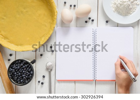 Rural white wood kitchen table with blank cooking book for recipe of blueberry pie. Pie ingredients (eggs, flour, dough, blueberries). Rustic background with space for shopping list