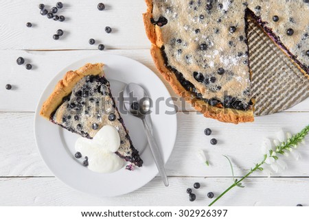 Blueberry pie on white wood kitchen table. Piece of pie on the plate served with cream and decorated with flower.