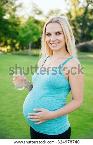 Pregnancy, sport and health lifestyle - happy pregnant woman drinking water from a glass in park in sunny summer day