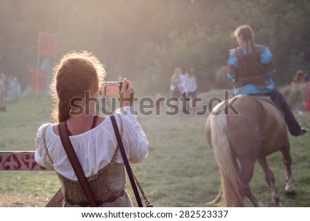 Girl wearing medieval clothes is taking a photo of a knight-girl on horseback.\
Urych, Ukraine - August 2: Tustan Medieval Culture Festival in Urych, Western Ukraine, on August 2, 2014