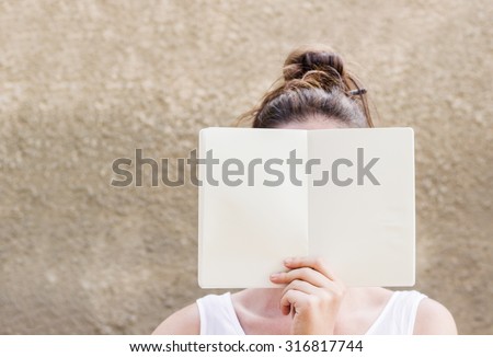 Close up Woman wearing white sleeveless blouse and pencil in her hair hiding her face behind empty white paper notebook, Emphasizing copy space, grey background