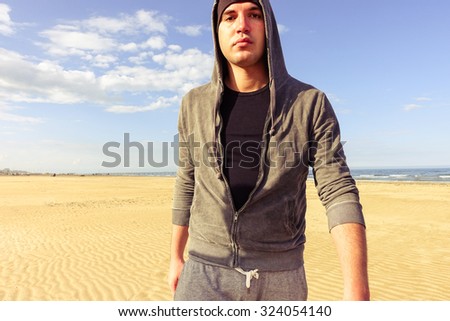 Young handsome tough man walking at beach - Attractive sporty hooded american guy having a break walk along ocean - Portrait of harsh face boxer training outdoor - Concept of people feelings and moods