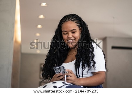 Happy young black woman in braids, white t-shirt, navy blue pants and earrings using her cellphone in her living room, with backlights Imagine de stoc © 