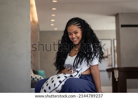 Happy young black woman in braids, white t-shirt, navy blue pants and earrings using her cellphone in her living room, with backlights Imagine de stoc © 