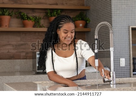 Cheerful young black brazilian woman with braided hair, white t-shirt, earrings and chain opening the faucet of her kitchen sink Imagine de stoc © 