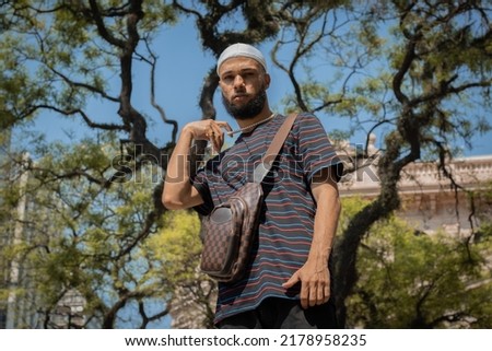 Young black brazilian man in durag, striped t-shirt, bag, ring, hands on his pearl chain in downtown São Paulo square Imagine de stoc © 
