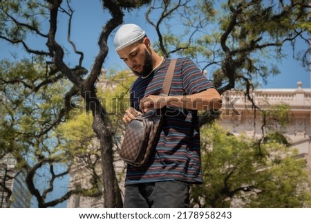 Young black brazilian man in durag, striped t-shirt, purse, ring, pearl chain opening her purse in downtown São Paulo square Imagine de stoc © 