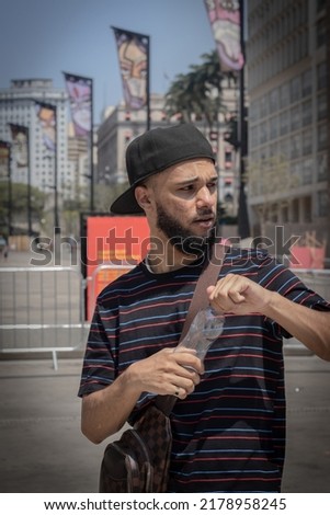 Young black brazilian man with beard, striped t-shirt, bag, cap, ring and pearl chain with bottle of water in his hands in downtown Sao Paulo Imagine de stoc © 