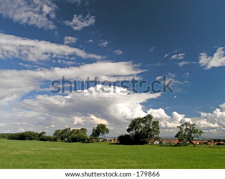 village behind trees under blue sky with small clouds and meadow