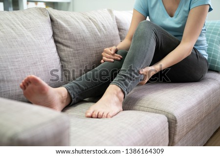 unrecognized young asian woman had pain at calves sitting on couch at home. chinese female in jeans hands massage legs on grey sofa. day off people sick illness painful body indoors in apartment. Imagine de stoc © 