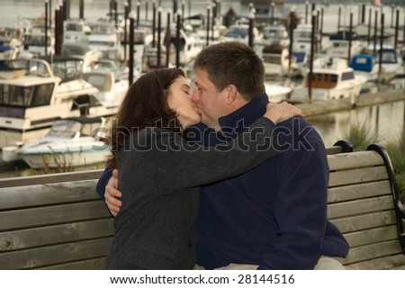 A couple kissing on a wooden bench in front of a boat marina.