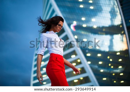 Attractive Business lady in red skirt and white shirt in front of glossy skyscraper with evening lights in windows. Woman wearing glasses walking in business-city with fresh wind in her hair.