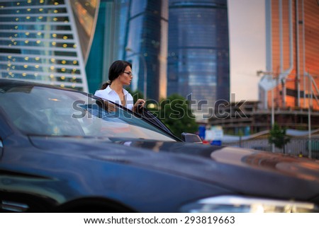 Business female going to drive a luxury car after work at evening time. Focus on her face and office buildings on background in a sunset light
