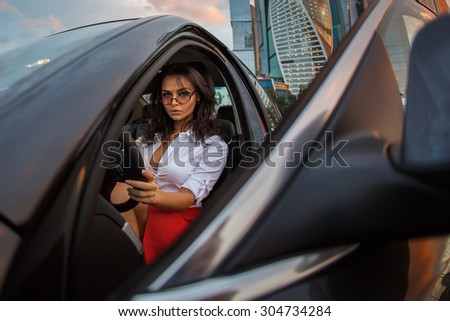 Business woman in a black car on drivers seat after hardworking day. A Brunette Lady sitting in a luxury vehicle looking from open door.