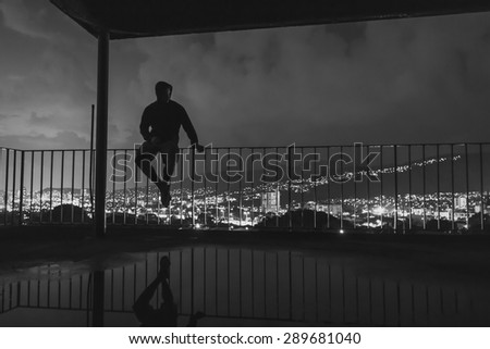 black and white of man sitting on railing overlooking city lights in the background at night.