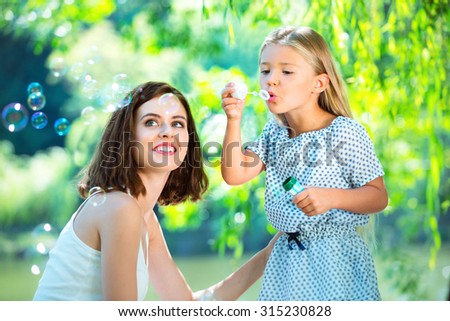 Smiling mother with daughter blowing bubbles. Smiling mother with daughter under the trees in the park. Rest time under the tree shadow. Enjoyment of blowing bubbles