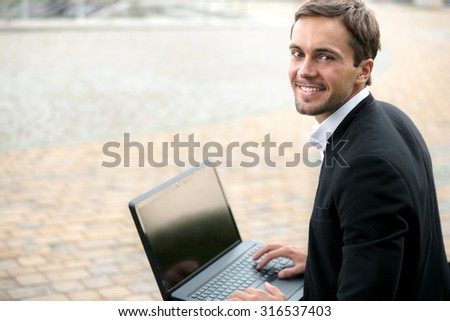 Young businessman is looking on us and smiling. He is holding his laptop on his legs. He is happy