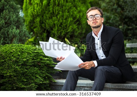 Serious man is sitting on the stairs and looking on us seriously. He is working with his notes. He wears dress code and glasses like businessman